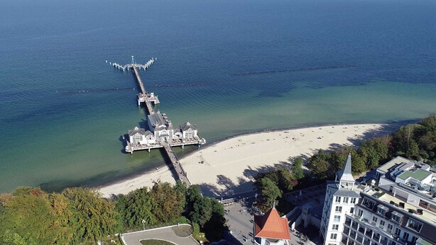 Vista aerea di una zona costiera con un molo che si protende nel mare, una spiaggia e edifici adiacenti.