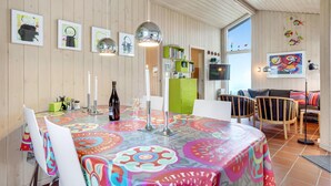A dining area with a colorful tablecloth, white chairs, and a bottle of wine. There are framed artworks on the wall and a green cabinet with books. - Amazing home in Sydals with WiFi (Sydals)