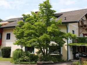 A building with a large tree in front, a balcony on the right, and a person sitting at a table outside.