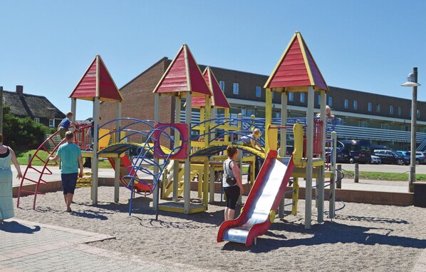 A playground with slides and climbing structures, surrounded by a sandy area and buildings in the background.