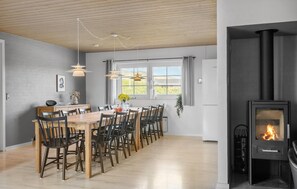 A modern dining area with a long wooden table, black chairs, and pendant lights. A black wood-burning stove is visible on the right. - Gorgeous home in Harboøre with sauna (Harboøre)