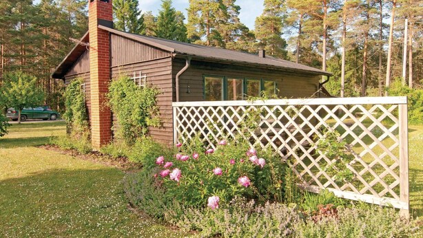 Eine kleine Holzhütte mit einem Backsteinsschornstein, umgeben von einem Garten mit rosa Blumen und einem weißen Gitterzaun.