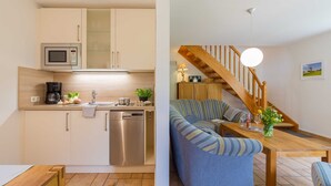 Une cuisine moderne équipée d’un four à micro-ondes, d’une machine à café et d’un lave-vaisselle. On y trouve une table à manger en bois, un canapé bleu et un escalier en bois avec une lampe suspendue.