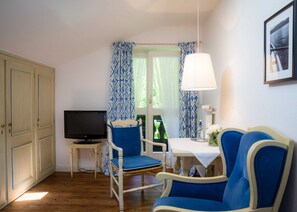 Una habitación con televisión, dos sillas azules, una mesa con lámpara y una ventana con cortinas estampadas.
