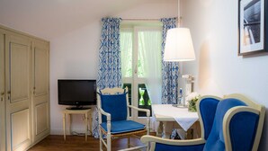 Una habitación con televisión, dos sillas azules, una mesa con lámpara y una ventana con cortinas estampadas.