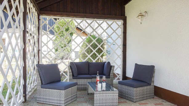 Uma área de varanda coberta com móveis de vime, incluindo um sofá e duas poltronas, e uma mesa de centro de vidro.