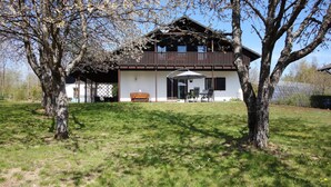 Uma casa de madeira de dois andares com uma varanda, cercada por árvores e um gramado.