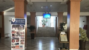 Vestíbulo de hotel con mostrador de recepción, una escalera y un letrero que dice 'Hocine Hotel'.