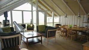 Een ruime kamer met houten plafond, grote ramen, een eettafel met stoelen en een bank met kussens.