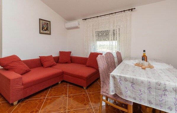 A living room with a red sectional sofa, a dining table with a floral tablecloth, and a window with lace curtains. - 1 bedroom awesome apartment in Betiga (Betiga)