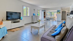 Amplia sala de estar con televisor, mobiliario blanco, comedor y cocina visible a través de una puerta abierta.
