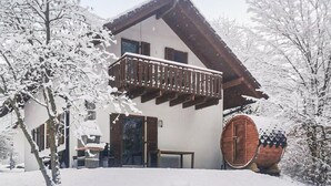 Una cabaña con balcón de madera, un barril de madera grande y árboles cubiertos de nieve.