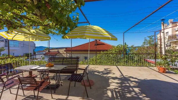 An outdoor patio with a table and chairs, two striped umbrellas, and a view of a building with a red roof.