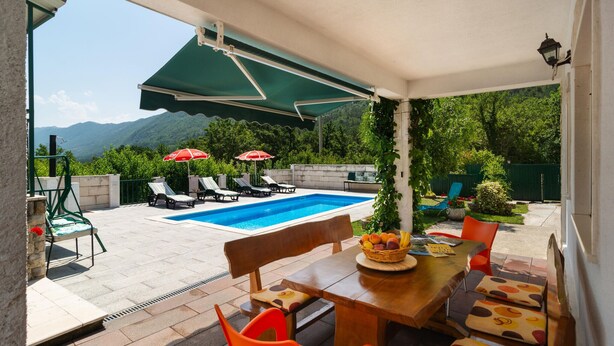 Zona junto a la piscina con piscina, sillas de descanso, sombrillas y una mesa con una cesta de fruta.