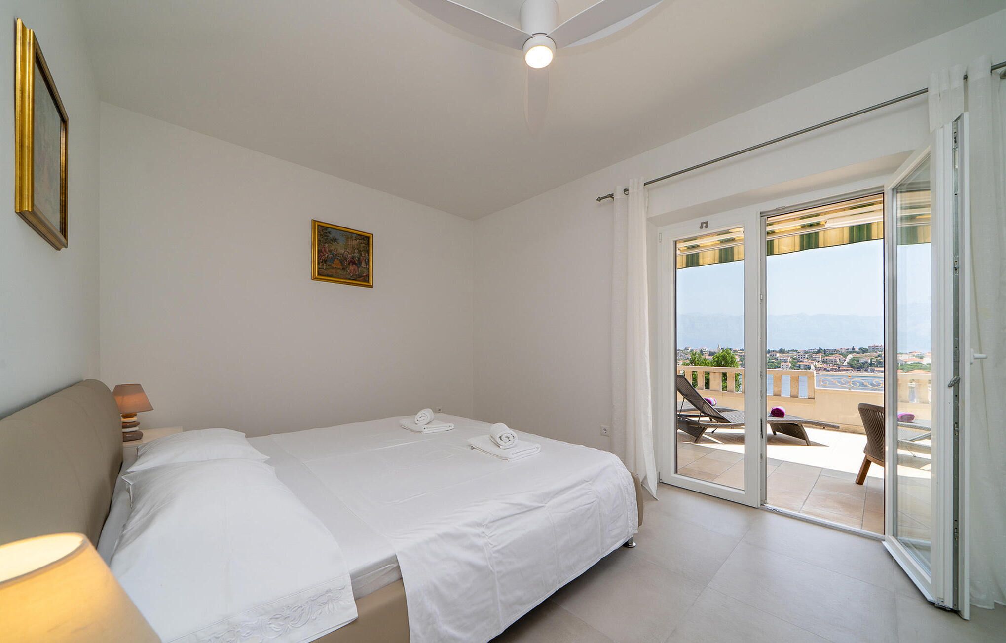 A hotel room with a bed, a painting, a lamp, and a balcony with chairs.