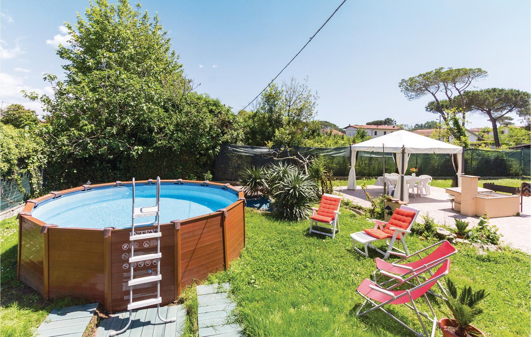 An outdoor swimming pool with a wooden deck, surrounded by greenery and a covered patio area with seating.