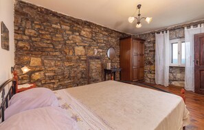 Une chambre avec un mur en pierre, un lit recouvert d’une couvre-lit à motifs floraux, une armoire en bois, un miroir et une fenêtre avec des rideaux.