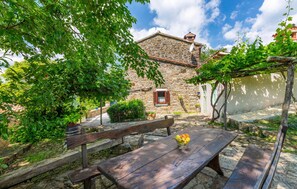 Un bâtiment rustique en pierre, doté d’une petite fenêtre, entouré d’une végétation luxuriante et agrémenté d’un banc en bois sur lequel se trouve un panier de fruits.