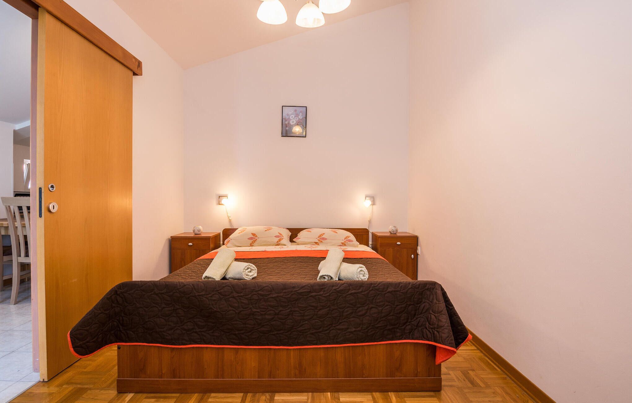 A neatly made bed with a brown quilt and orange accents, two bedside tables with lamps, a wooden door, and a small framed picture on the wall.