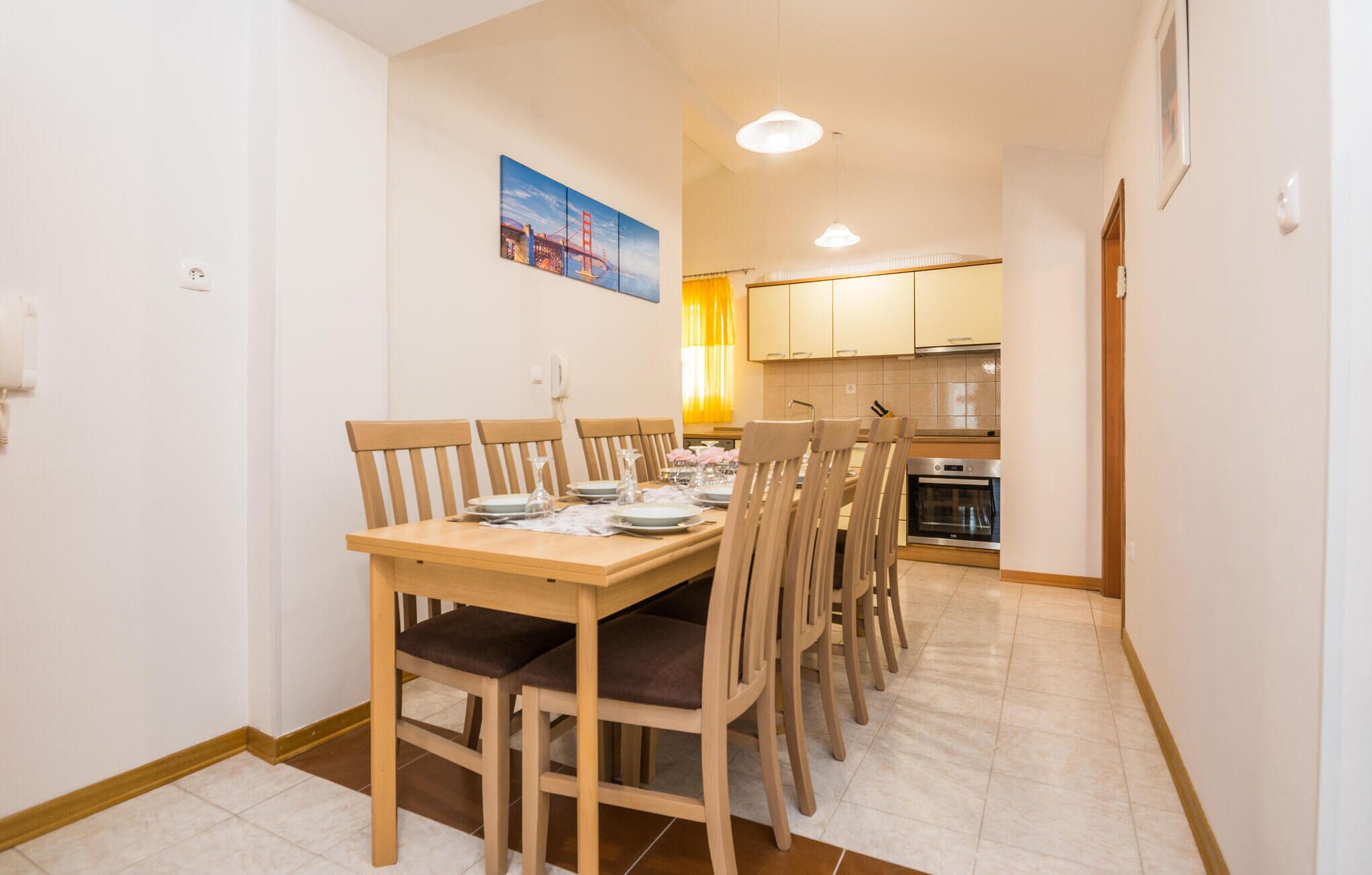 A dining area with a wooden table and chairs, a kitchen in the background, and a painting on the wall.