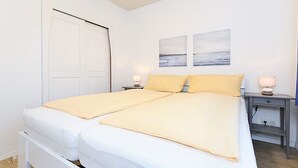 Een tweepersoonsbed met wit beddengoed en een gele deken, een nachtkastje met lamp, en een wandplank.