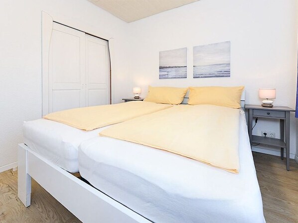 Un lit double avec une literie blanche et une couverture jaune, une table de chevet avec une lampe et une étagère fixée au mur.