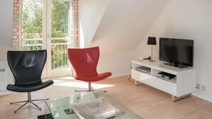 A modern living room with a glass coffee table, a black and a red chair, a white entertainment unit with a TV, and a large window with curtains. - Gorgeous home in Blåvand with sauna (Blåvand)
