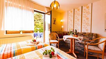 A hotel room with two beds, a sofa, a small table, and a view of greenery outside.