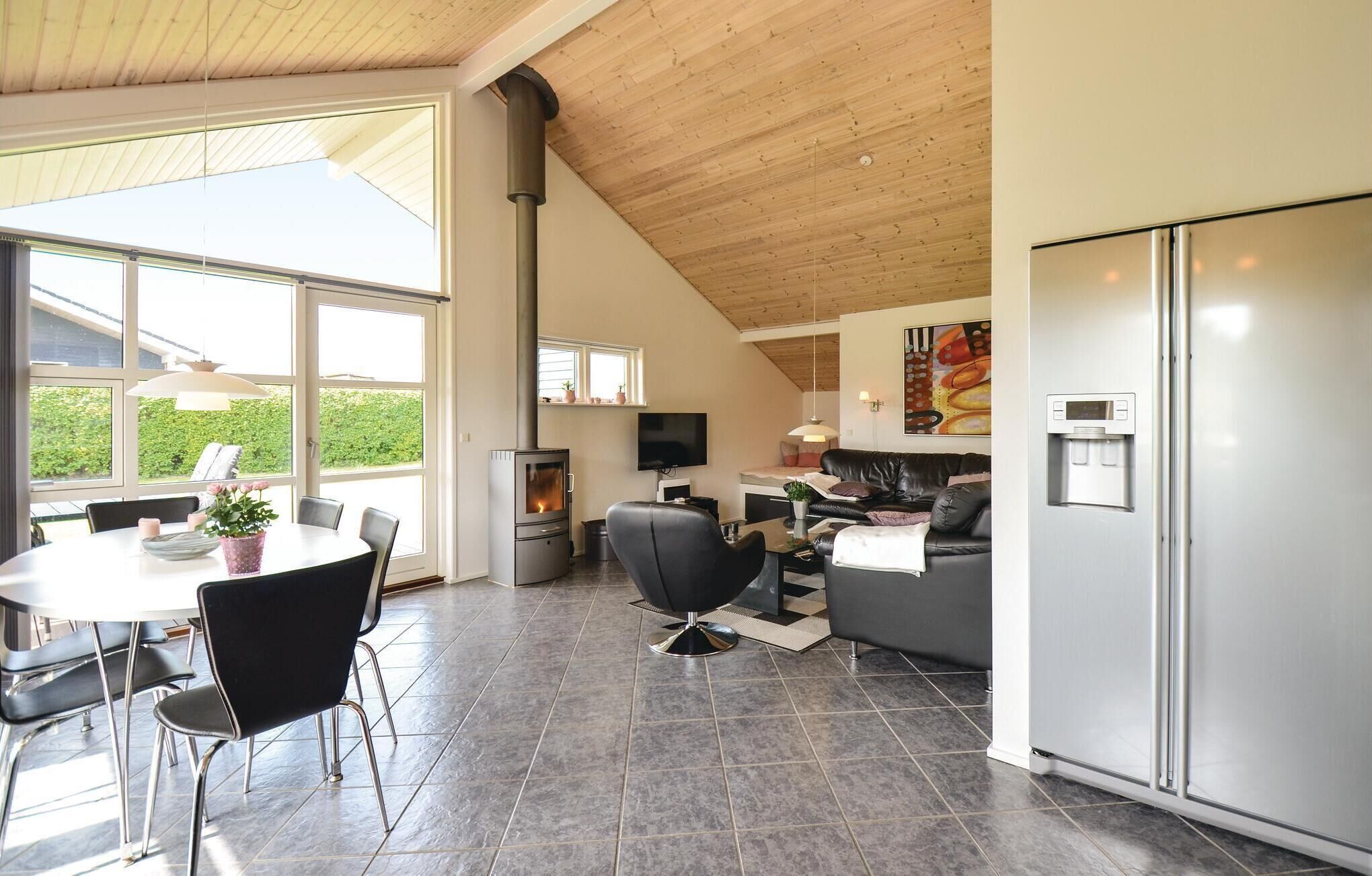 A modern kitchen with a built-in stove, a dining area with a table and chairs, a sofa, and a large window offering a view of greenery.