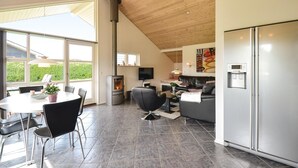 A modern kitchen with a built-in stove, a dining area with a table and chairs, a sofa, and a large window offering a view of greenery. - Stunning home in Knebel with sauna (Knebel)
