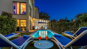 A poolside area with lounge chairs, a small table with two glasses of red drink, and a building with large windows and a covered patio. - Stunning home in Malinska with sauna (Malinska)