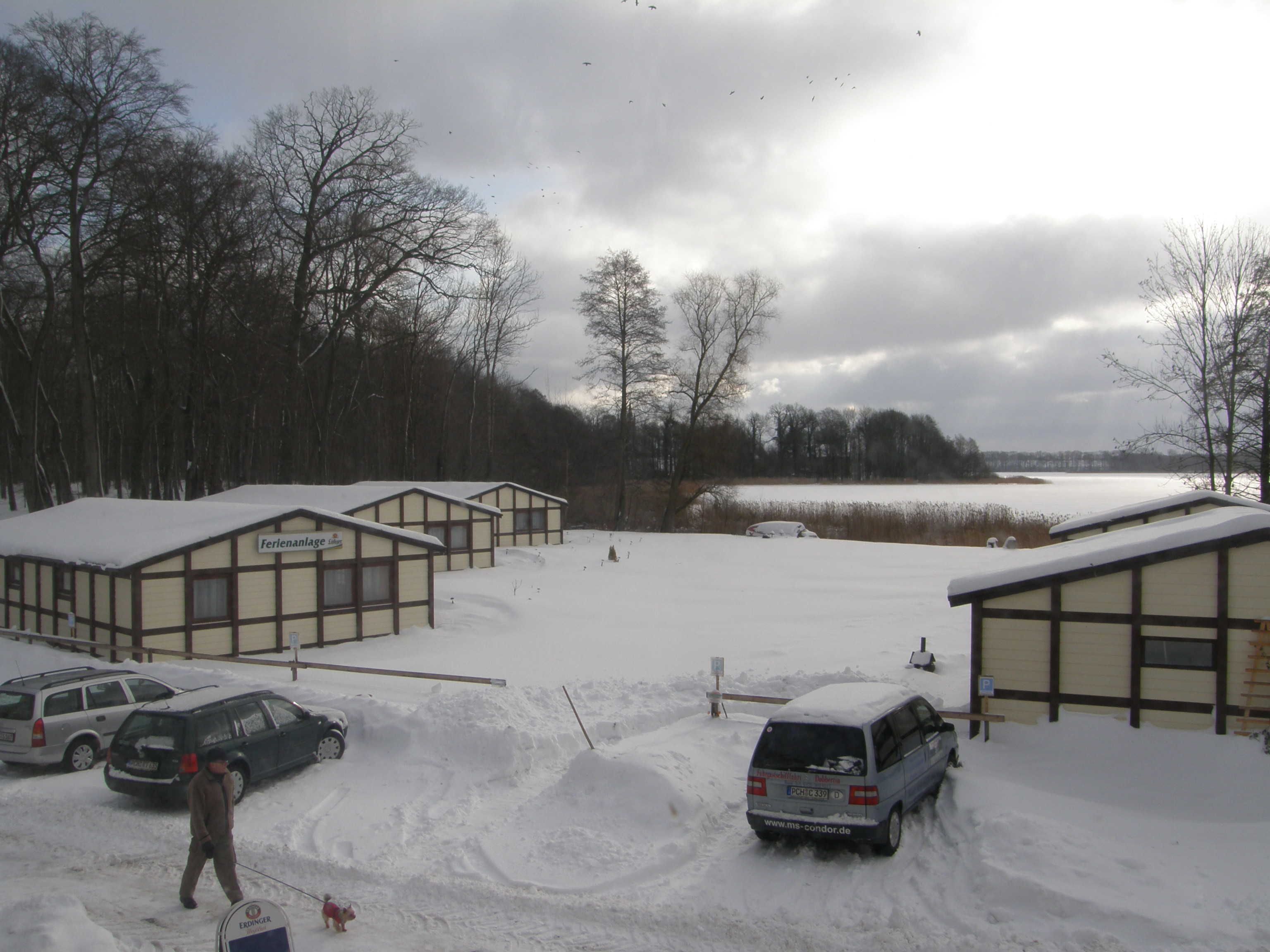 Una hilera de cabañas sobre un terreno cubierto de nieve, una masa de agua al fondo y un cielo nublado.