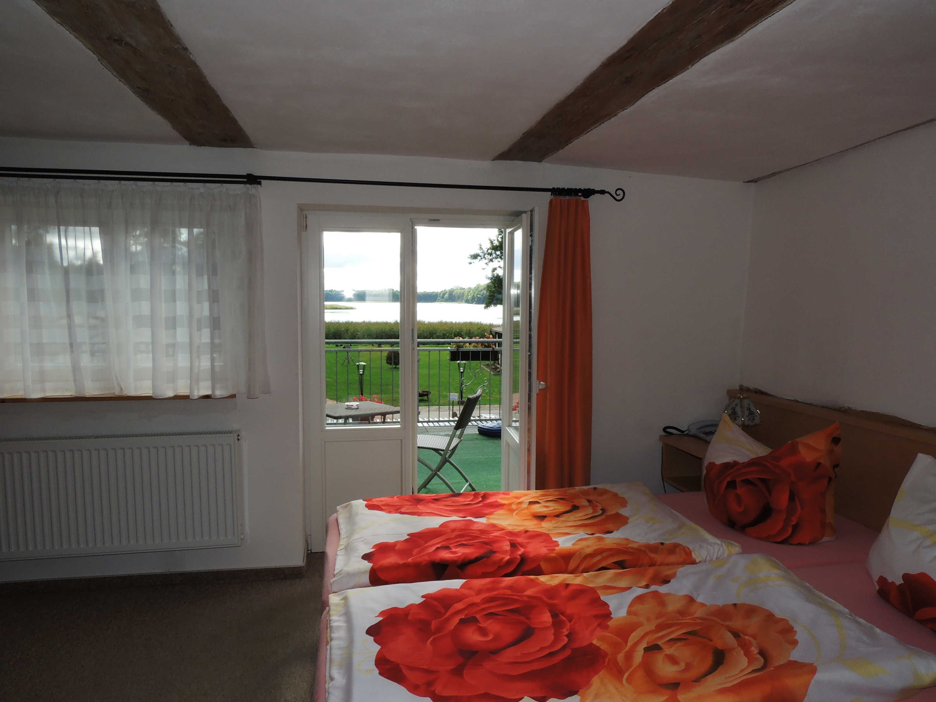 A bedroom with a bed featuring a floral bedspread, a window with sheer curtains, and a view of a green outdoor area.