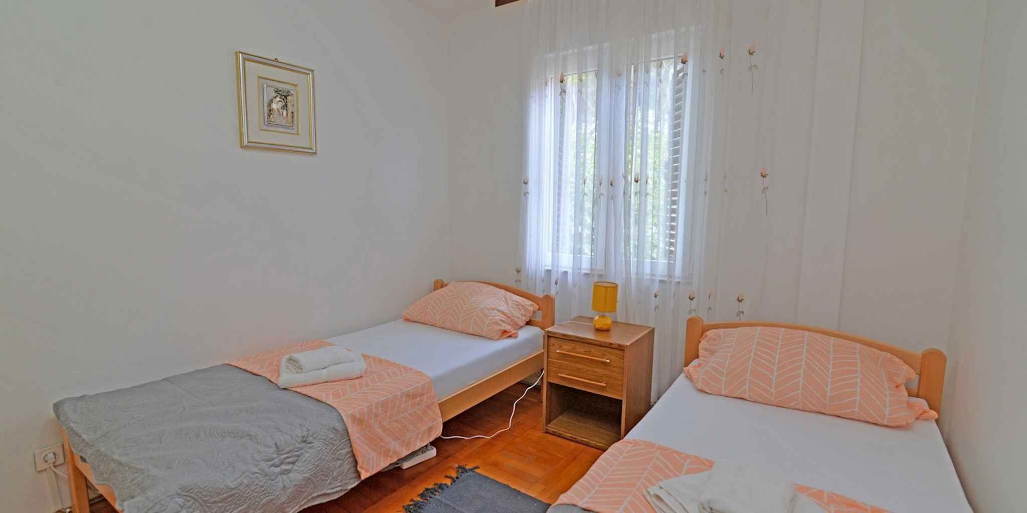 Une chambre avec deux lits simples, une table de chevet en bois, un bureau et une fenêtre avec des rideaux légers.