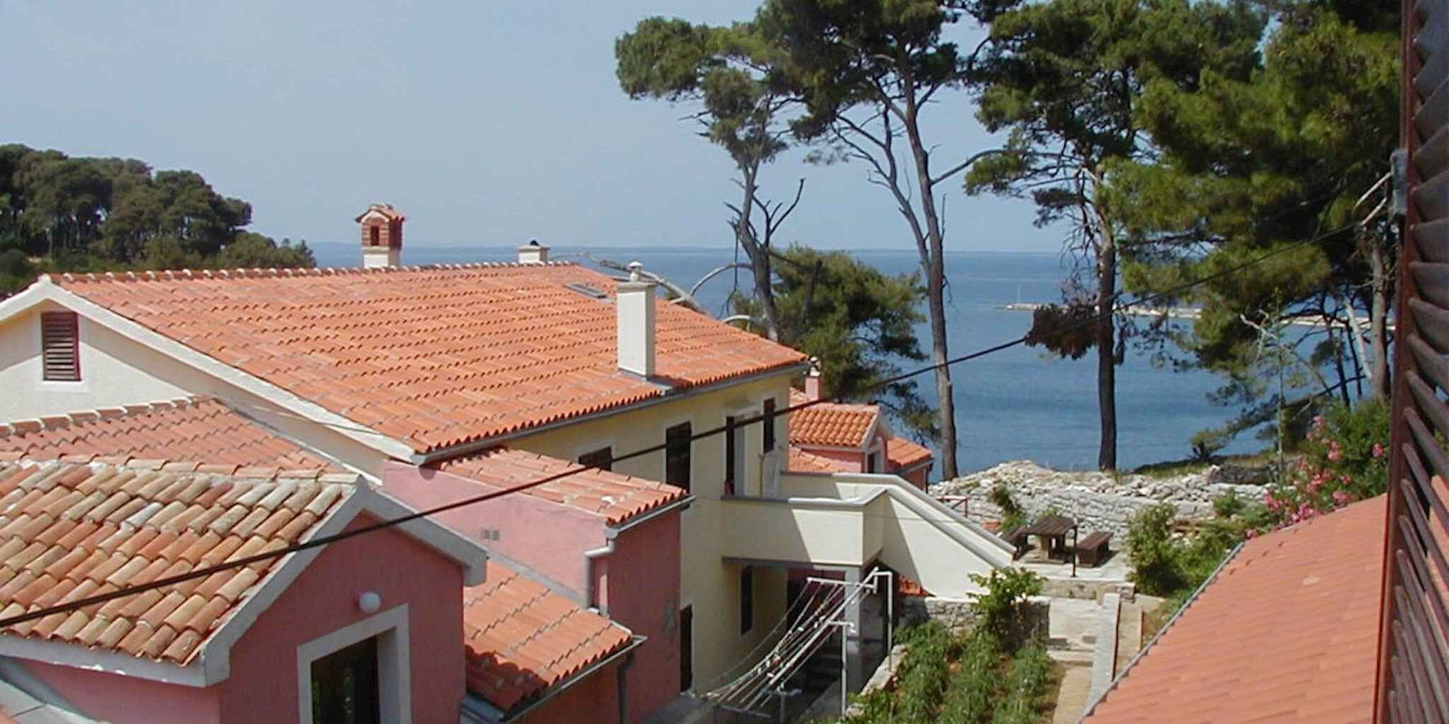 A coastal residential area with terracotta-tiled roofs, a clear view of the sea, and a few trees.