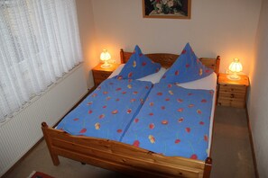A bed with a blue floral bedspread, two pillows, two bedside lamps, a wooden headboard, a wooden nightstand, and a window with white lace curtains. - Vacation apartment Bianca - House Reese (Clausthal-Zellerfeld)
