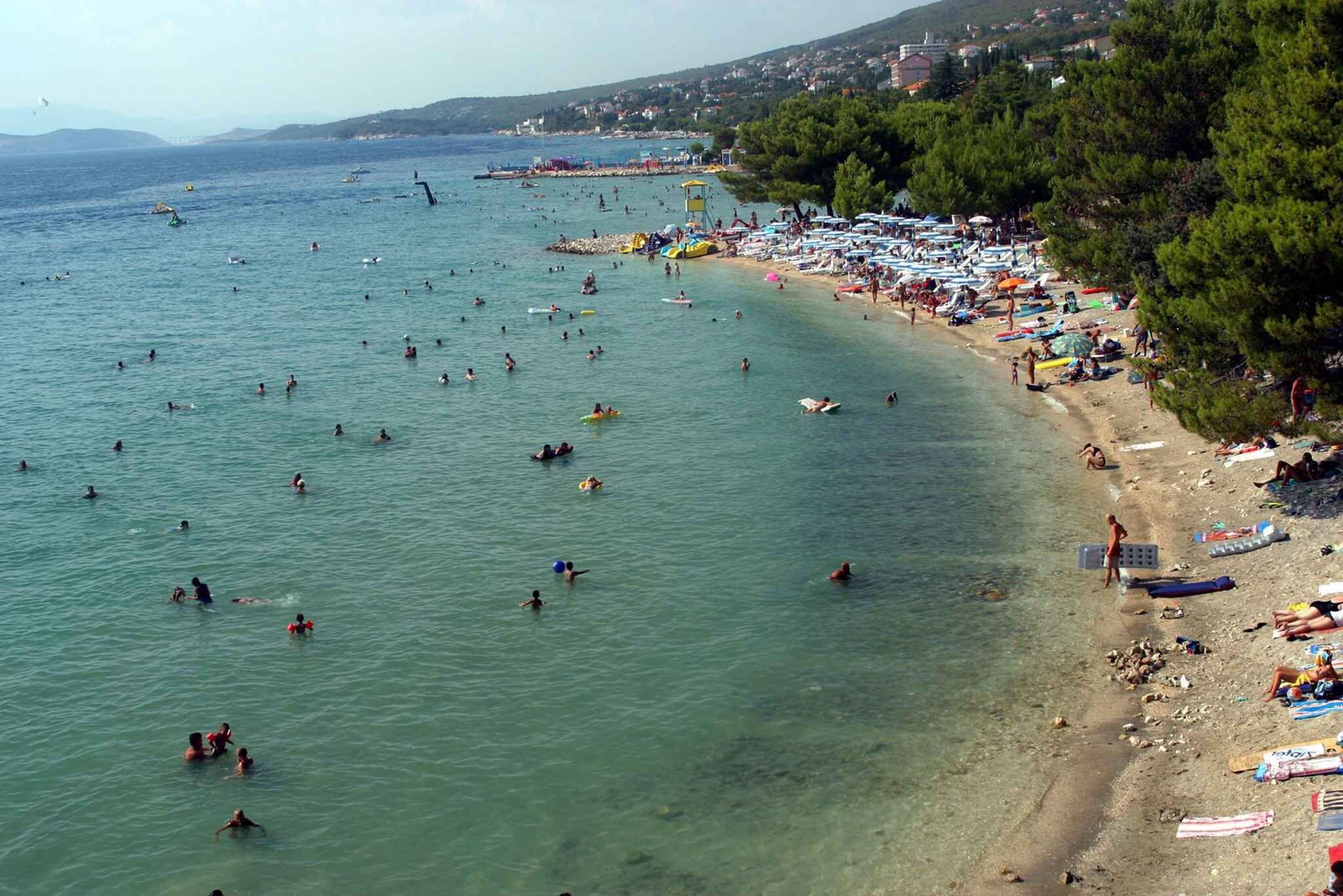 Een strand met helder water, mensen die zwemmen en zonnebaden, en een rij parasols aan de kust.