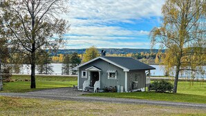 A small, single-story cabin with a porch, situated in a grassy area with trees and a lake in the background. - 2 bedroom awesome home in Svanskog (Svanskog)