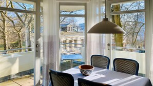 Un comedor con una mesa y sillas, ventanas amplias con cortinas y vistas a un edificio y árboles al exterior.
