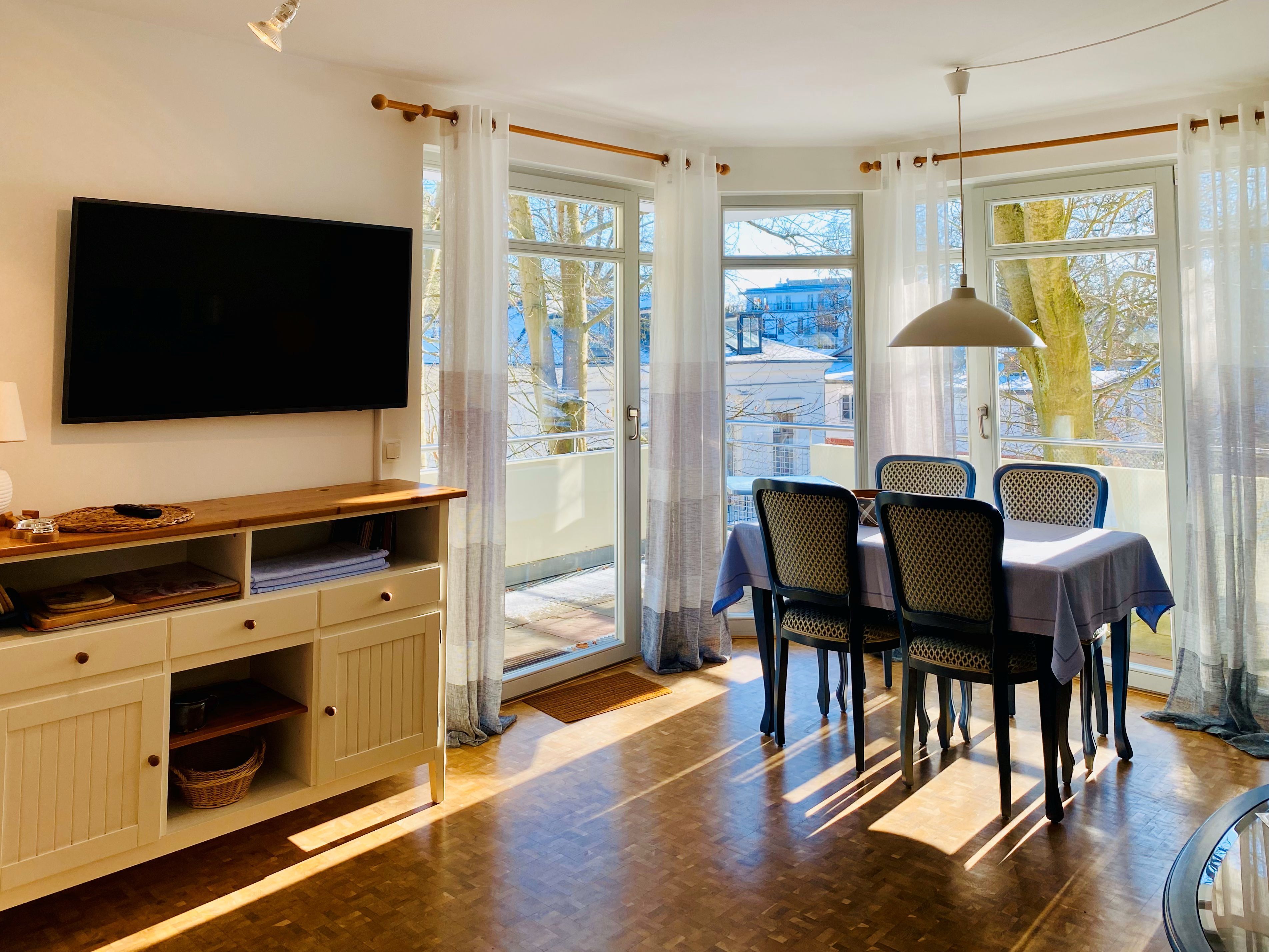 Un coin repas avec une table et des chaises, un téléviseur fixé au mur et une vue sur l’extérieur grâce à de grandes fenêtres.