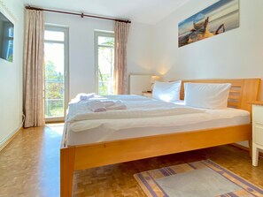 Ein helles Schlafzimmer mit einem hölzernen Bett, weißer Bettwäsche und Blick ins Freie durch große Fenster.
