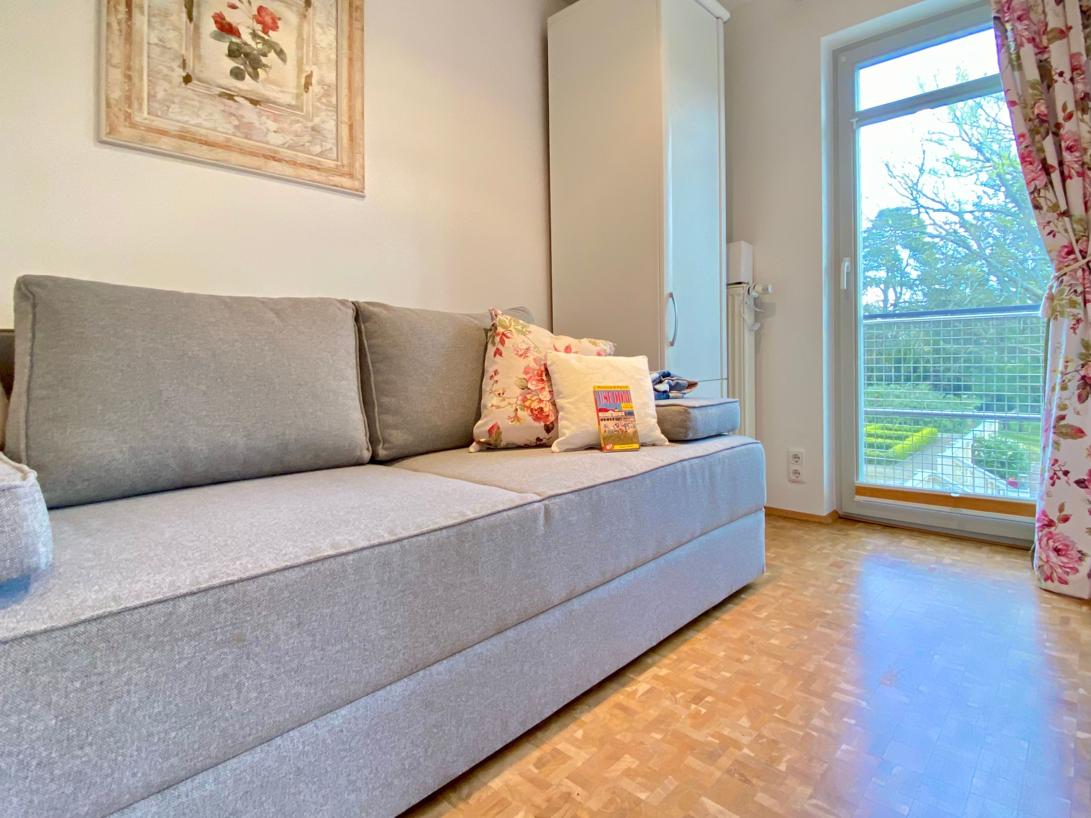 A grey sofa with floral cushions and a magazine, a framed picture on the wall, and a view of greenery through a window with a curtain.