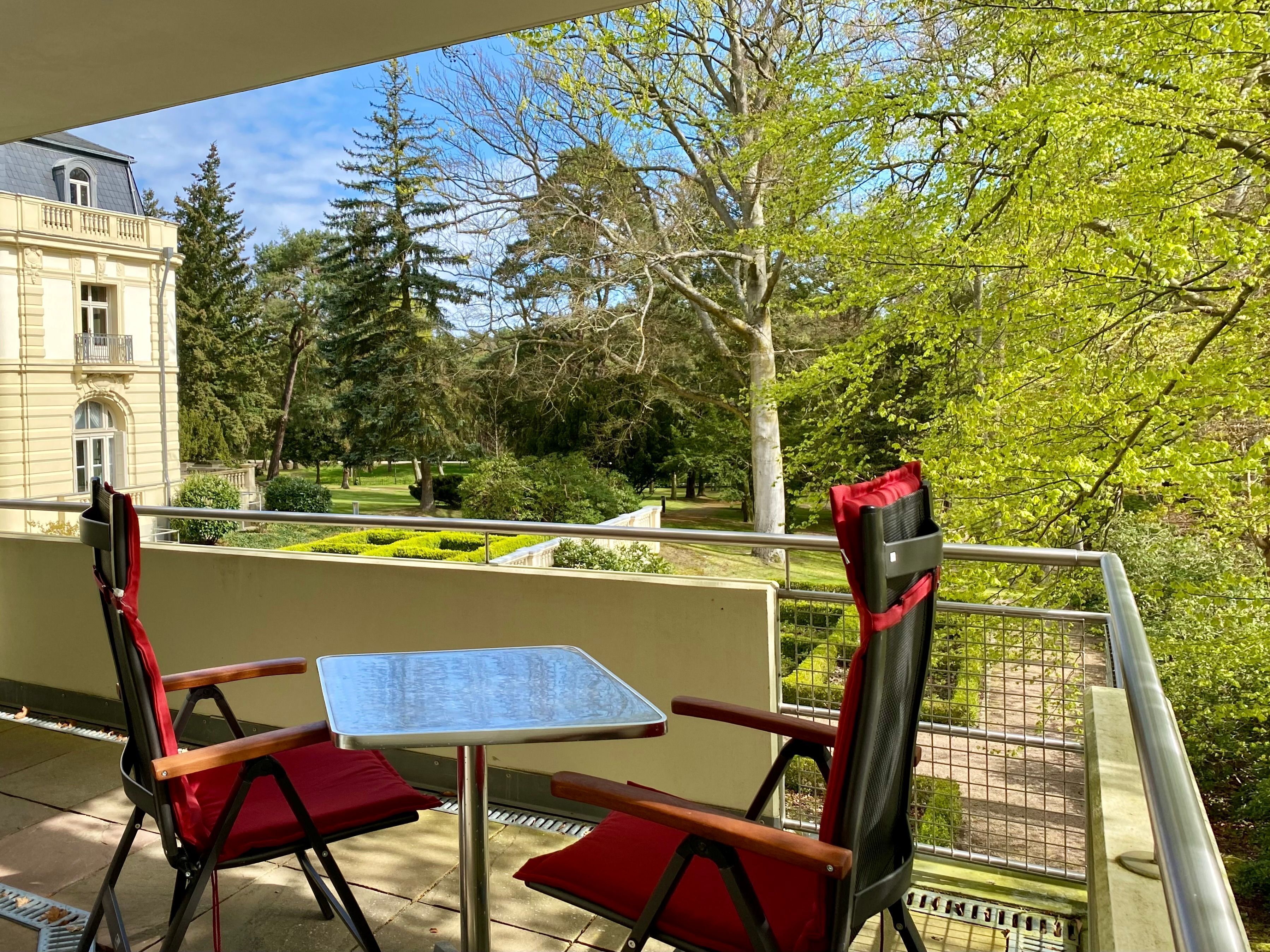 Un balcón con dos sillas rojas y una pequeña mesa, con vistas a un jardín cuidado y un edificio histórico.