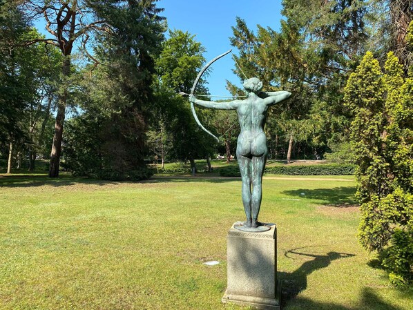 A statue of a figure with an arrow, set on a pedestal in a park with trees and grass.