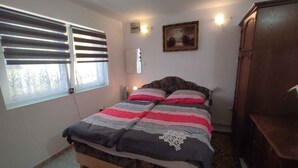 A bedroom with a bed, a window with blinds, a nightstand, a lamp, a framed picture on the wall, and a wooden cabinet. - Studio with covered terrace and billiards, near the harbour (Balatonboglár)
