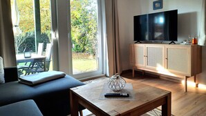 Ein Wohnzimmer mit einer Couch, einem Couchtisch, einem Fernseher auf einem Holzschrank und Blick auf einen Garten durch große Fenster.