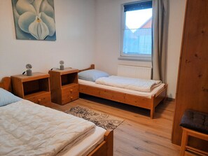 A small, cozy bedroom with two single beds, a wooden dresser, a nightstand, a window with a view, and a radiator.