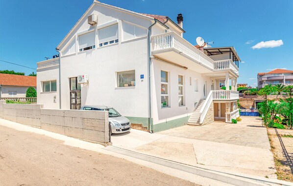 Ett vitt hus i två våningar med en balkong, en bil parkerad framför och en liten trädgård.