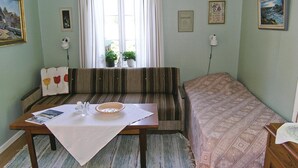 A room with a striped sofa, a small table with a placemat, a bed with a patterned bedspread, a window with curtains, and framed pictures on the walls.