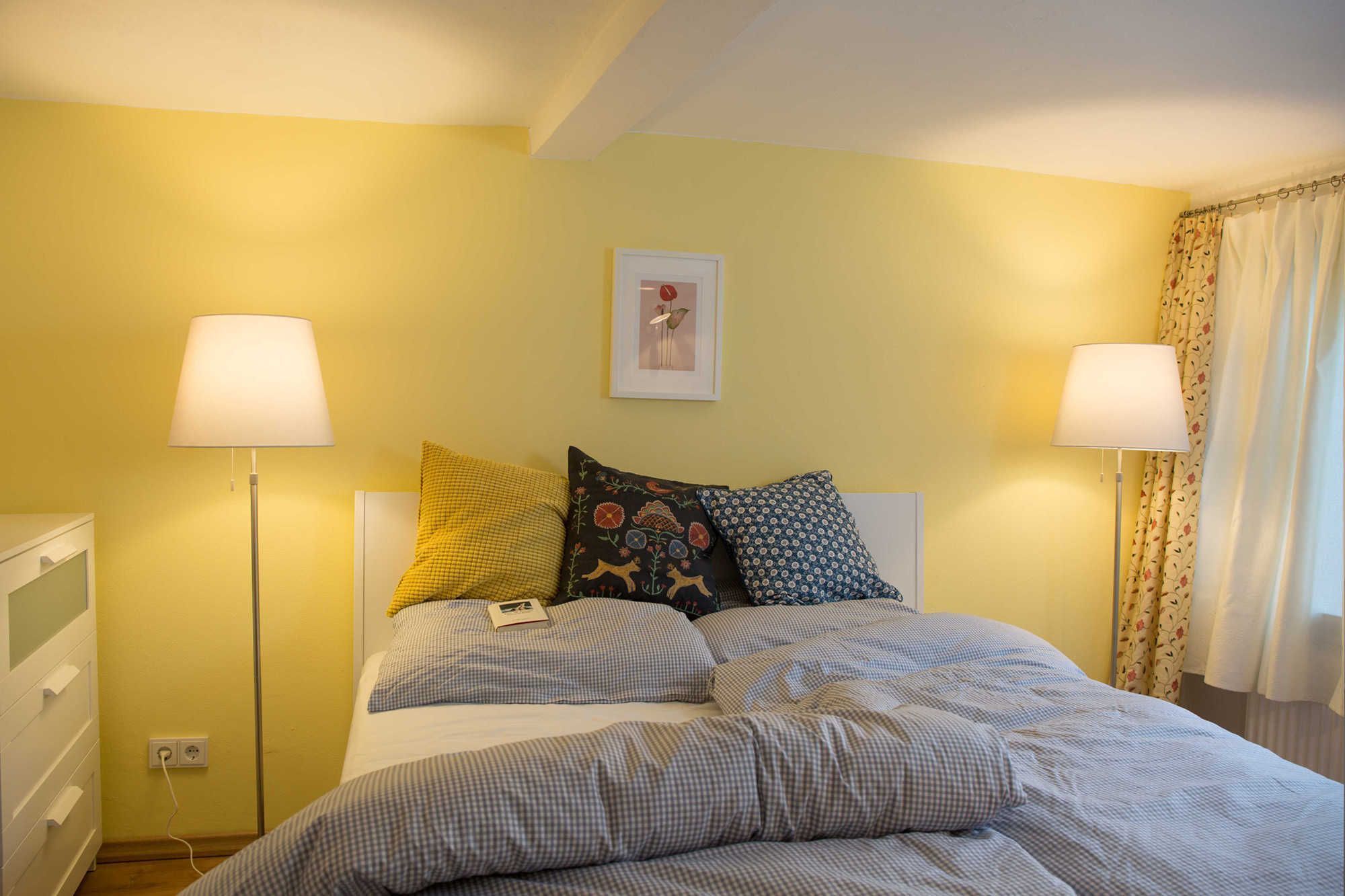 A bed with checkered bedding, two bedside lamps, a framed picture on the wall, and a window with curtains.